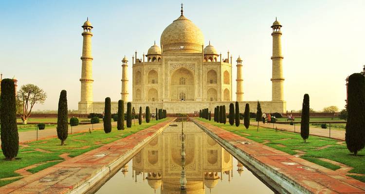 Vue de face du Taj Mahal se reflétant dans le long bassin, encadrée par des jardins entretenus dans une lumière matinale chaleureuse.