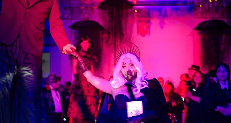 Un échassier guide une femme costumée en vampire à travers un hall vivement éclairé.