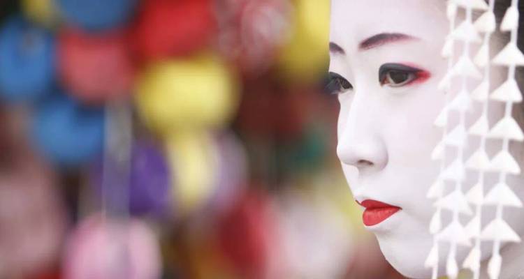 Retrato de cerca de una geisha con maquillaje blanco y labios rojos sobre un fondo borroso y colorido.