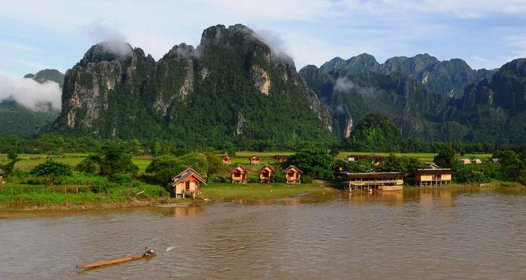 A rural riverside scene with houses and mountains under a clear sky.