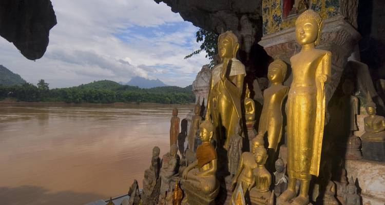 Golden Buddha statues in a cave overlooking a river and hills.