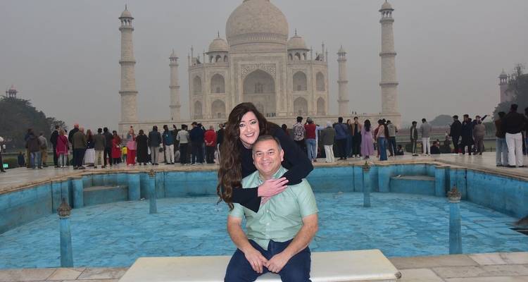 Couple poses affectionately on bench in front of iconic Taj Mahal reflected pool on hazy day.
