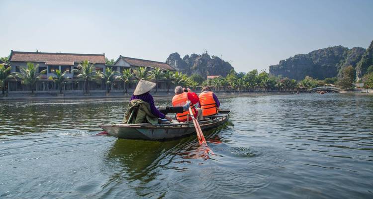 People in a small boat rowing on a wide river with lush surroundings.