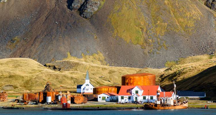 Station baleinière abandonnée avec église blanche, réservoirs de couleur rouille et bâtiments aux toits rouges situés contre une pente de montagne rocheuse et abrupte à côté d'une baie calme.