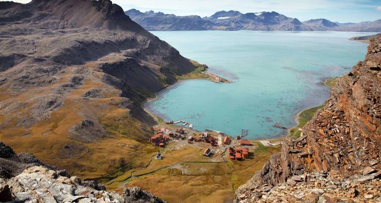 Vue panoramique élevée d'un fjord turquoise entouré de montagnes escarpées et d'une petite agglomération aux teintes rouille sur ses rives.