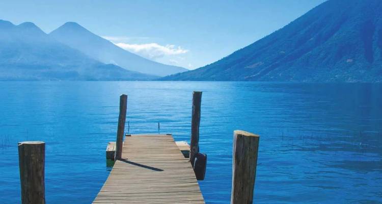 Un muelle de madera que se extiende hacia un lago azul tranquilo, rodeado por montañas volcánicas lejanas bajo un cielo despejado.