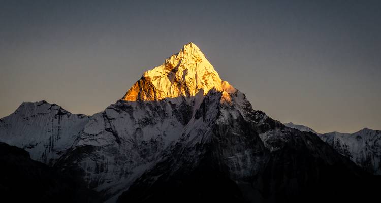 Un pico del Himalaya cubierto de nieve brillando con la cálida luz del amanecer contra un cielo oscuro y despejado.