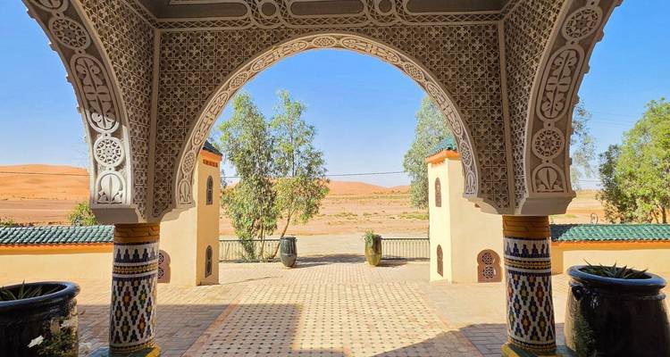 Un ornado arco marroquí enmarca la vista de dunas del desierto y árboles de eucalipto bajo un cielo azul despejado.