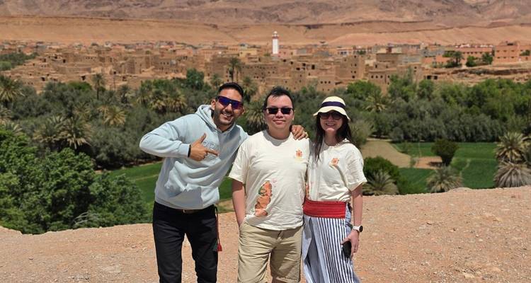 El guía y dos viajeros sonríen con un amplio pueblo oasis y palmeras extendiéndose detrás de ellos.