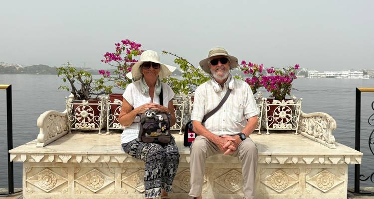 Un couple est assis sur un banc en marbre sculpté orné de bougainvillées, dominant un lac tranquille.