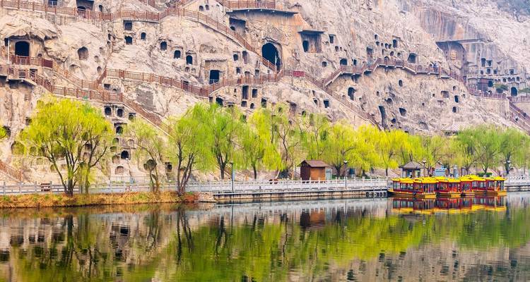Les grottes de Longmen creusées dans la falaise se reflétant dans une rivière calme avec des arbres printaniers et des bateaux de tourisme