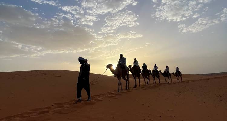 Gruppe von Menschen auf Kamelen, die bei Sonnenaufgang über Sanddünen wandert.