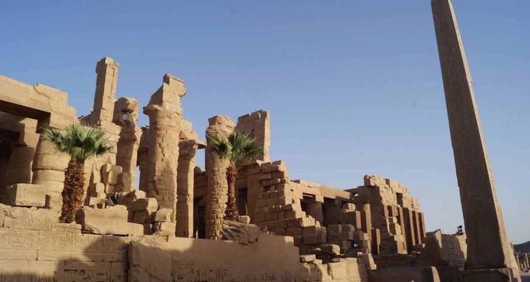 Ruinas antiguas con palmeras y un obelisco bajo un cielo despejado.