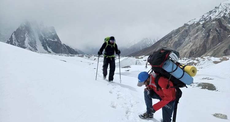 Twee klimmers met zware rugzakken en wandelstokken banen zich een weg over een besneeuwde pas bij bewolkte omstandigheden.