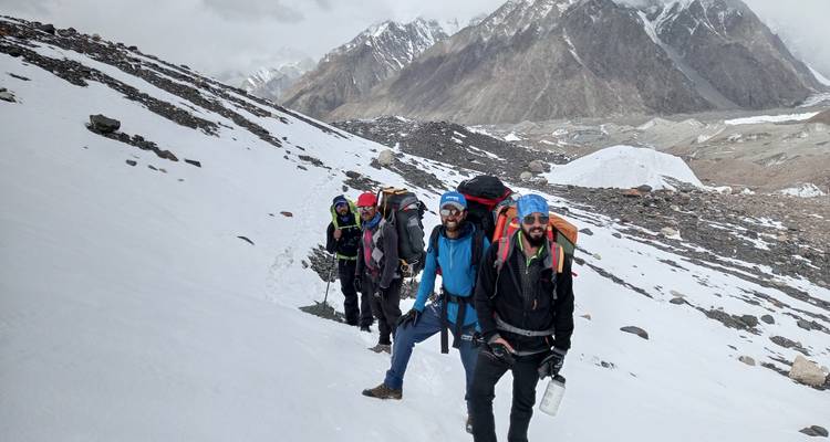 Una línea de excursionistas escala una pendiente empinada cubierta de nieve con imponentes picos rocosos detrás.