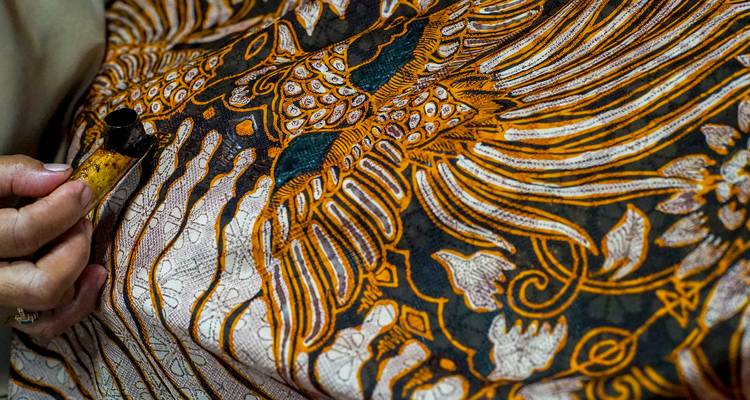 Primer plano de una mano aplicando cera a una tela batik de patrones ricos en tonos naranjas, dorados y verde azulado.