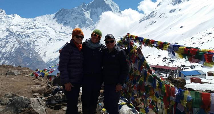 Three people standing in front of prayer flags and snowy mountains.