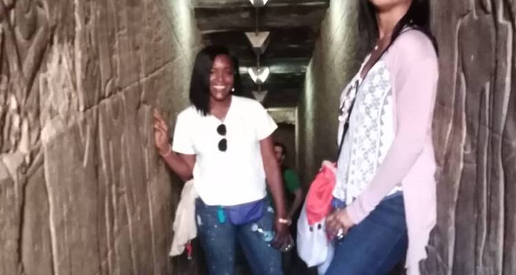Dos mujeres posan dentro de un estrecho pasaje de piedra antigua con paredes talladas e iluminación tenue.
