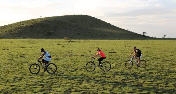 Three cyclists riding across a grassy plain with a hill in the background.