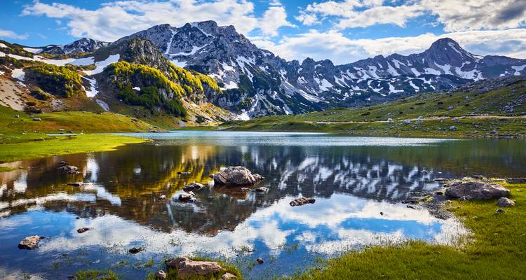Lac alpin tranquille reflétant des pics escarpés striés de neige et des pentes herbeuses sous un ciel bleu partiellement nuageux.