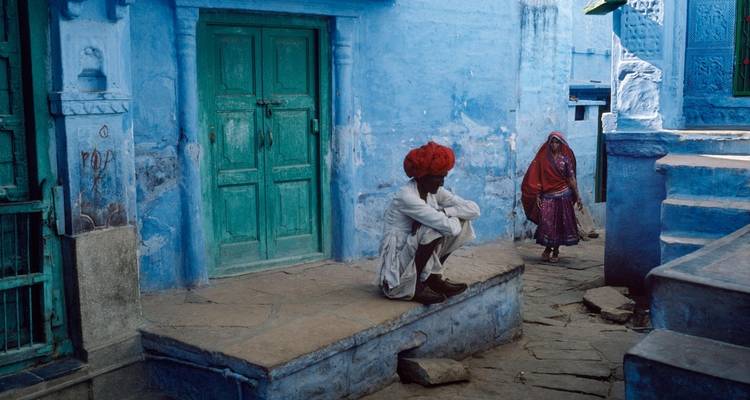 Hombre con turbante rojo sentado con una mujer en sari azul en una calle azul.
