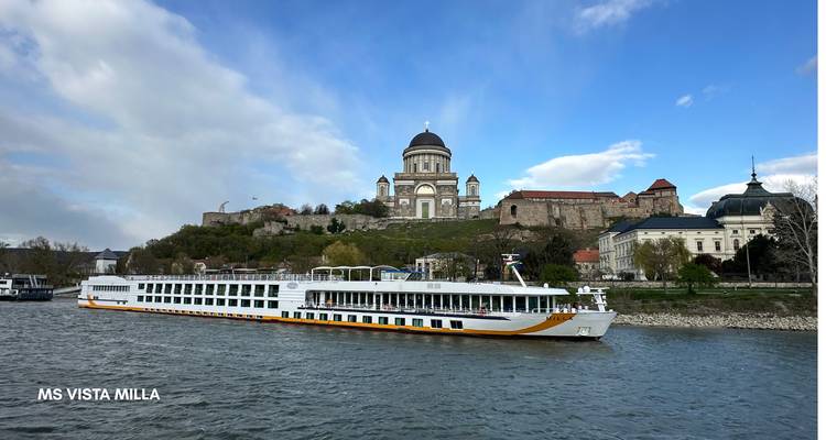 Ein Flusskreuzfahrtschiff, das an einem historischen Gebäude unter blauem Himmel vorbeifährt.