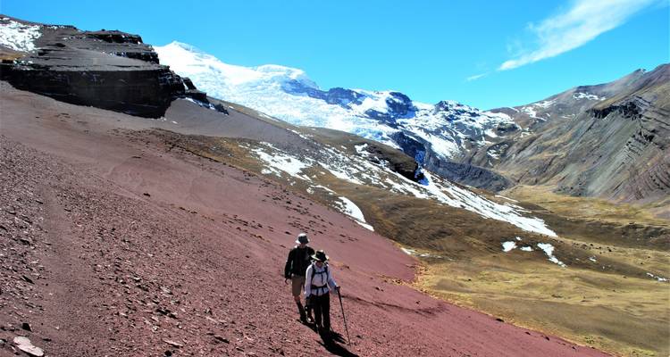 Twee wandelaars die lopen op een rood bergpad met besneeuwde toppen in de verte.