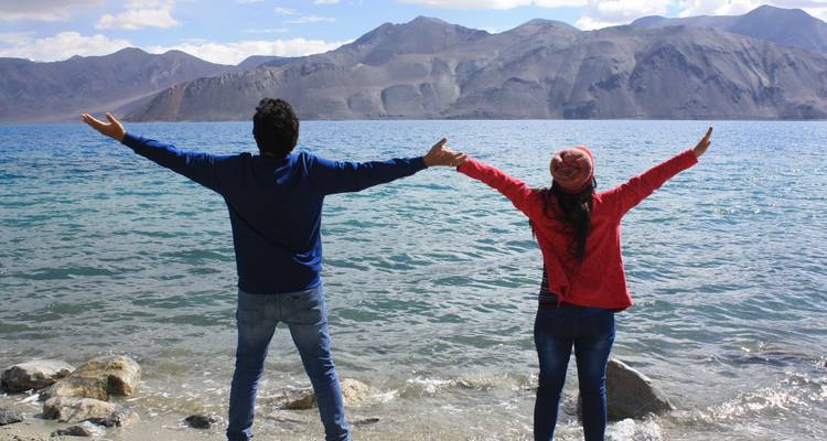 Dos personas paradas junto a la orilla de un lago con los brazos extendidos.