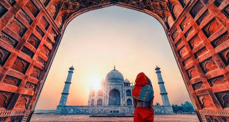 Zonsopgang uitzicht op de Taj Mahal omlijst door een imposante rode zandsteen boog met een reiziger in een sari die ernaar kijkt.