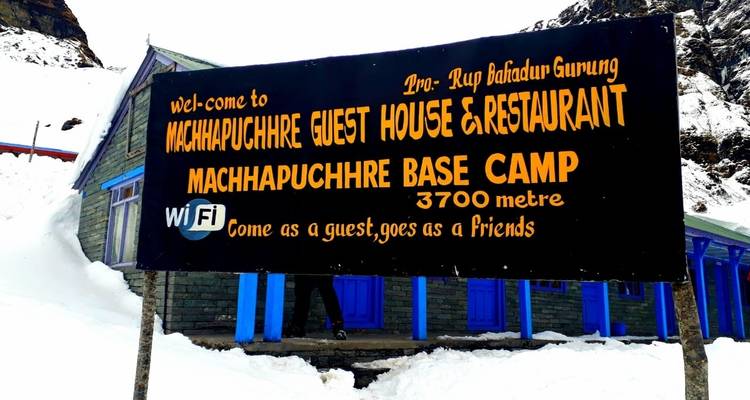 Schild für Machhapuchhre Gästehaus und Restaurant mit Schnee und Gebäude im Hintergrund.