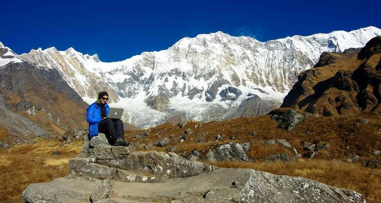 Een persoon die een laptop gebruikt, zittend op rotsen met besneeuwde bergen op de achtergrond.