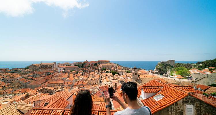 Zwei Menschen genießen den Blick auf eine historische Stadt mit Terrakotta-Dächern am Meer.