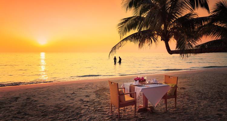 Ambiente romántico de playa con mesa y sillas, palmera, y atardecer sobre el océano.