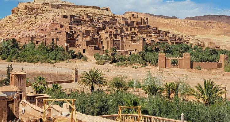 Vista expansiva del pueblo fortificado de arcilla de Ait Benhaddou situado sobre un lecho de río con exuberantes palmeras en primer plano y colinas áridas detrás.