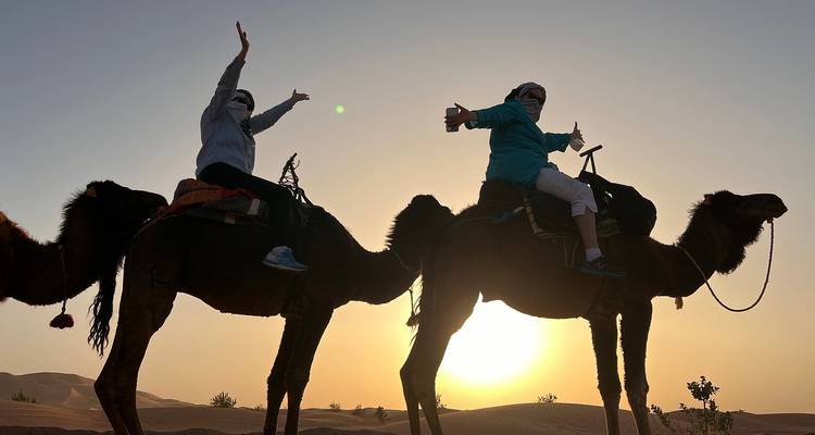 Dos viajeros en camellos posan alegremente mientras el sol naciente los siluetea contra las doradas arenas del desierto.