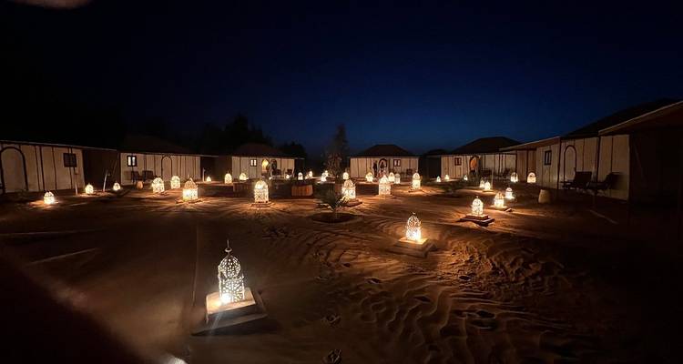 Patio de campamento del desierto por la noche iluminado por docenas de linternas ornamentadas que proyectan luz cálida sobre la arena ondulada entre tiendas de lona.