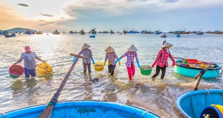 Mujeres caminando en el agua al amanecer con barcos pesqueros de fondo.