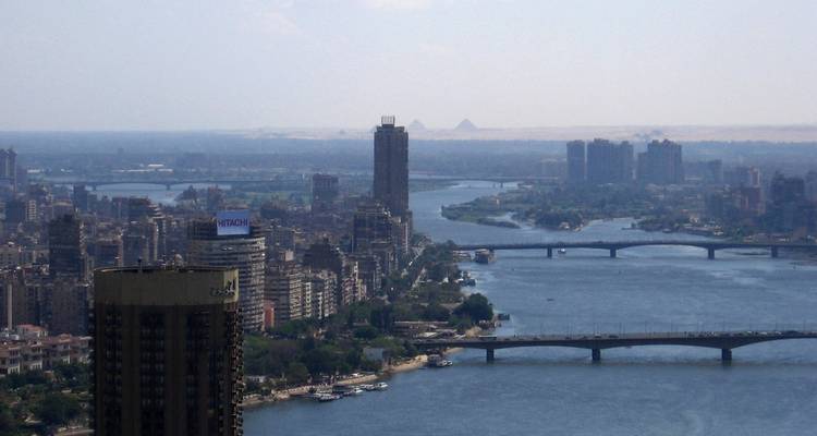Luftaufnahme einer Stadt mit einem Fluss, hohen Gebäuden und Pyramiden im Hintergrund.