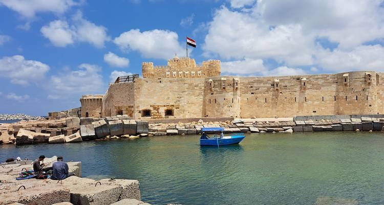 A fortress by the sea with a boat and people relaxing nearby.