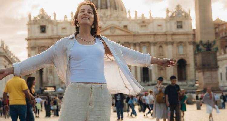 Une voyageuse joyeuse écarte largement les bras sur la place Saint-Pierre avec la basilique doucement floue en arrière-plan.
