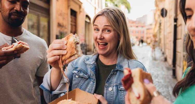 Une femme montre avec espièglerie un sandwich dégoulinant pendant que des amis goûtent la nourriture de rue dans une ruelle romaine.