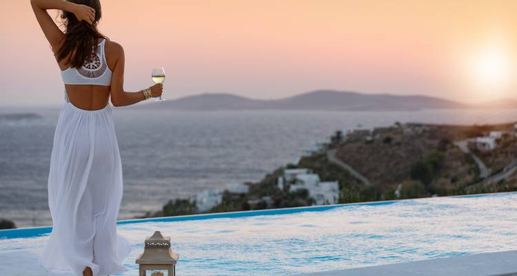 Mujer disfrutando de una bebida junto a una piscina infinita con vista al mar al atardecer.
