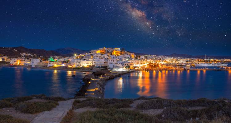 Nachtansicht von Naxos mit Stadtlichtern, die sich unter einem sternenklaren Himmel auf dem Wasser spiegeln.