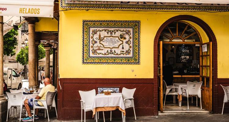 Una fachada de café amarillo brillante con mesas al aire libre donde un comensal solitario se sienta bajo un colorido letrero de mapa callejero de cerámica.