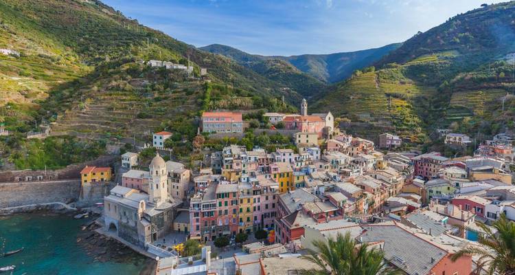 Un village coloré à flanc de falaise avec des maisons aux couleurs pastel qui dégringolent vers un petit port, entouré de collines verdoyantes en terrasses.
