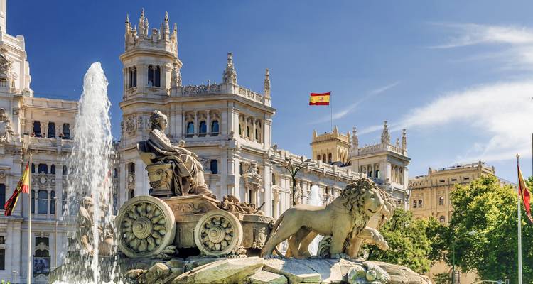 Grande fontaine avec des lions de pierre et une figure de déesse devant un palais orné couronné par le drapeau espagnol par une journée ensoleillée et lumineuse.