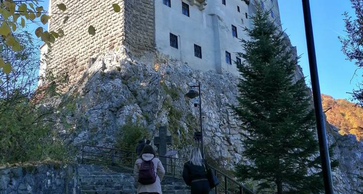 Deux visiteurs gravissent des marches de pierre vers les imposants murs du château de Bran perché sur la falaise au milieu du feuillage automnal