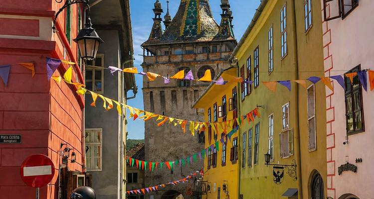 Colorful medieval street hung with bunting leading to Sighisoara’s clock tower under clear blue skies.
