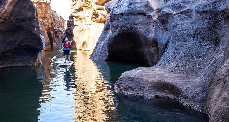 Pratiquant de stand-up paddle naviguant dans une gorge étroite aux parois hautes avec une eau calme et réfléchissante.