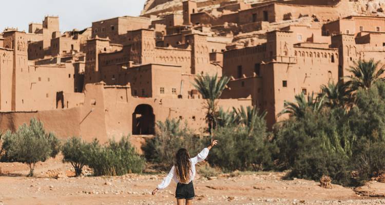 Person with arms raised towards adobe buildings in a desert setting.
